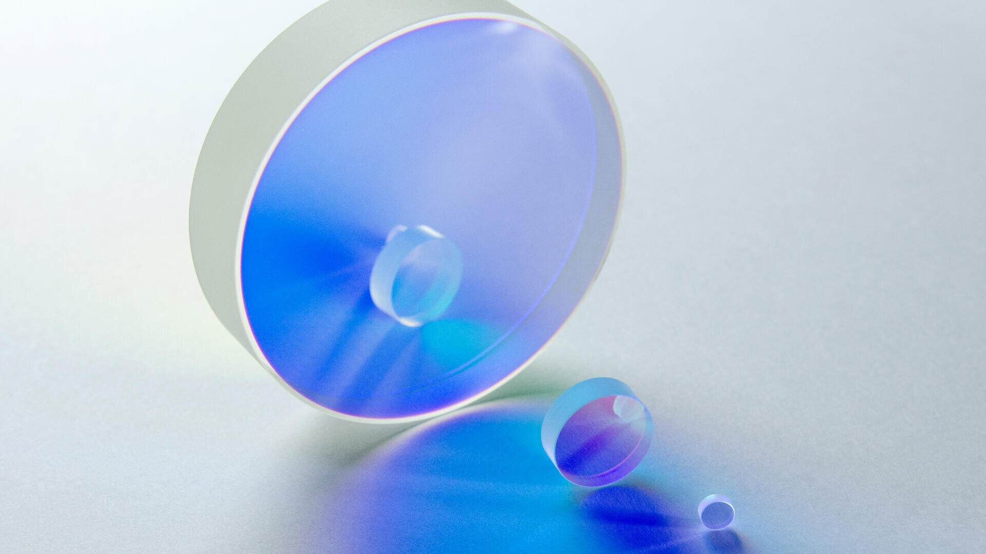 A large round optical lens and two smaller glass lenses rest on a pale surface, casting blue and purple light reflections and shadows.