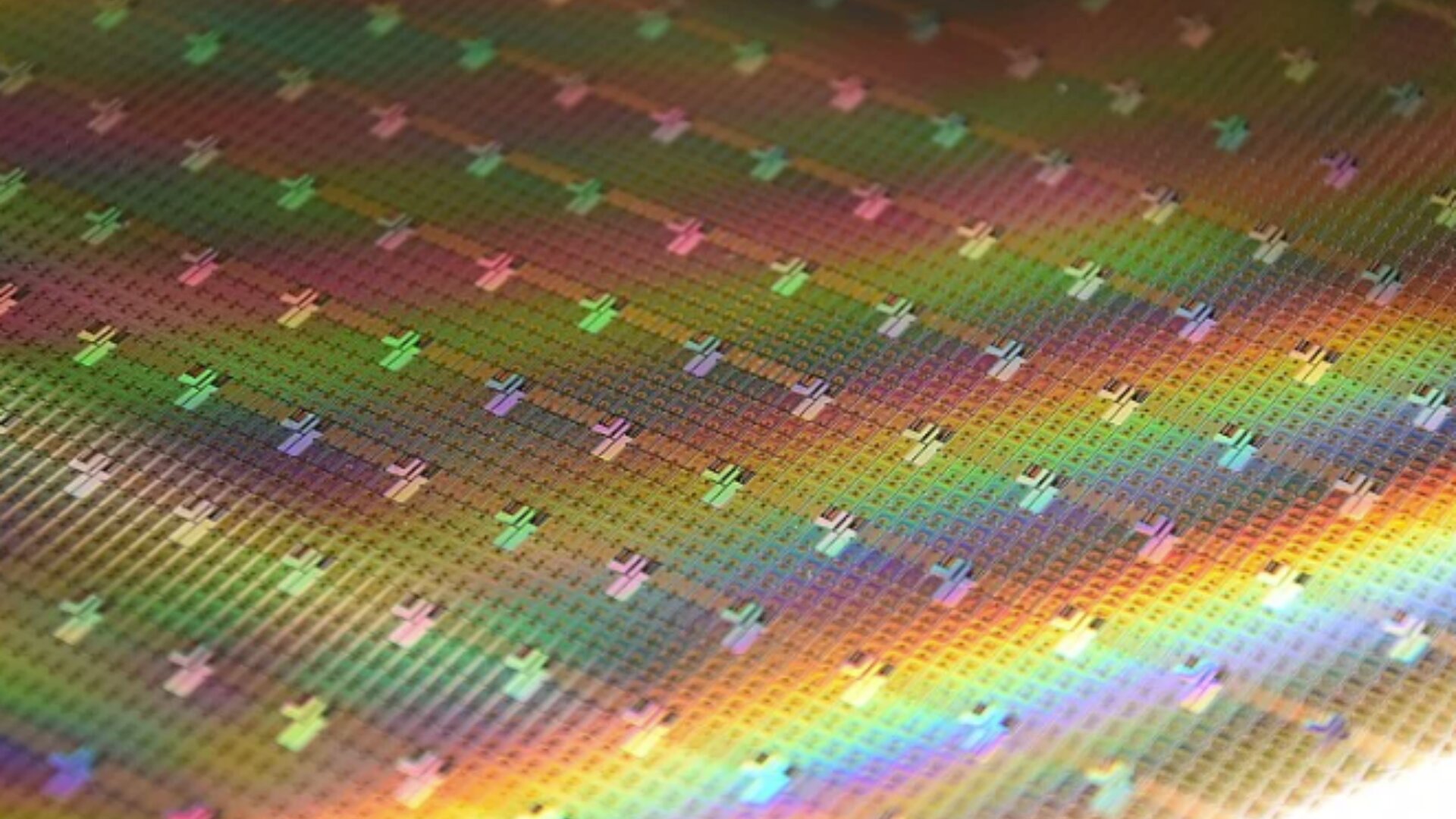 A close-up of a silicon wafer with microchips, displaying a grid of small, square chips reflecting iridescent rainbow colors.