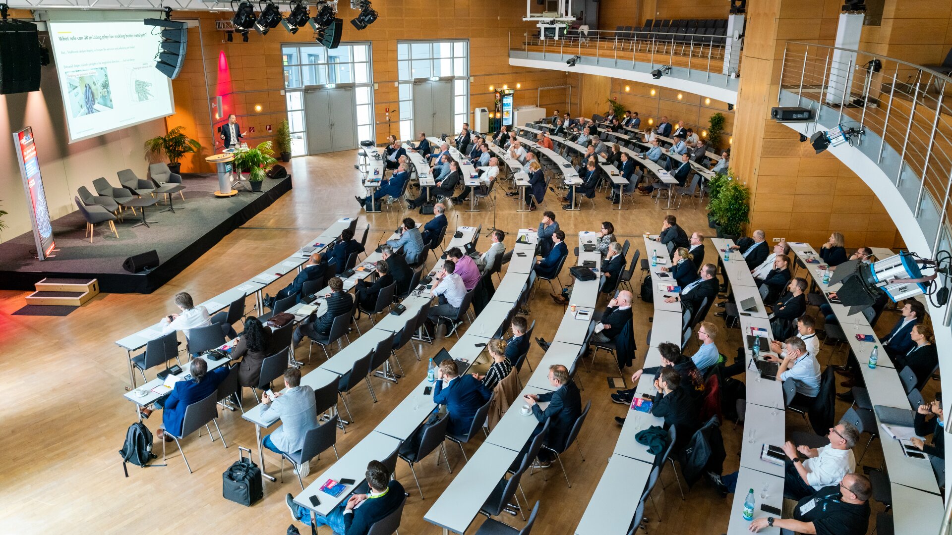 Eine große Gruppe von Menschen sitzt an langen Tischreihen in einem modernen Hörsaal und hört einem Redner zu, der Folien auf einer Leinwand im vorderen Teil des Raumes präsentiert. Einige Teilnehmer machen sich Notizen oder schauen auf ihre Laptops.