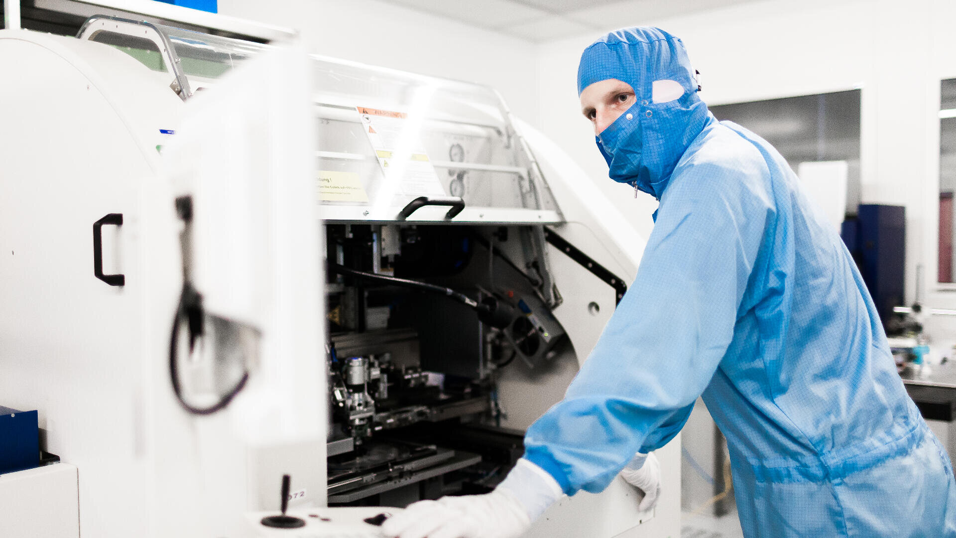 Eine Person, die einen blauen Reinraumanzug, eine Maske und Handschuhe trägt, bedient Maschinen in einem sterilen Labor oder einer Produktionsumgebung.