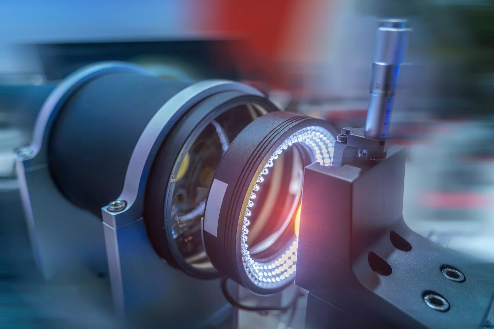 Nahaufnahme eines optischen Instruments mit einer Linse, umgeben von einem Ring aus LED-Leuchten, montiert auf einem Metallrahmen. Es wird möglicherweise in der wissenschaftlichen oder industriellen Bildgebung oder Inspektion verwendet. Der Hintergrund ist unscharf.