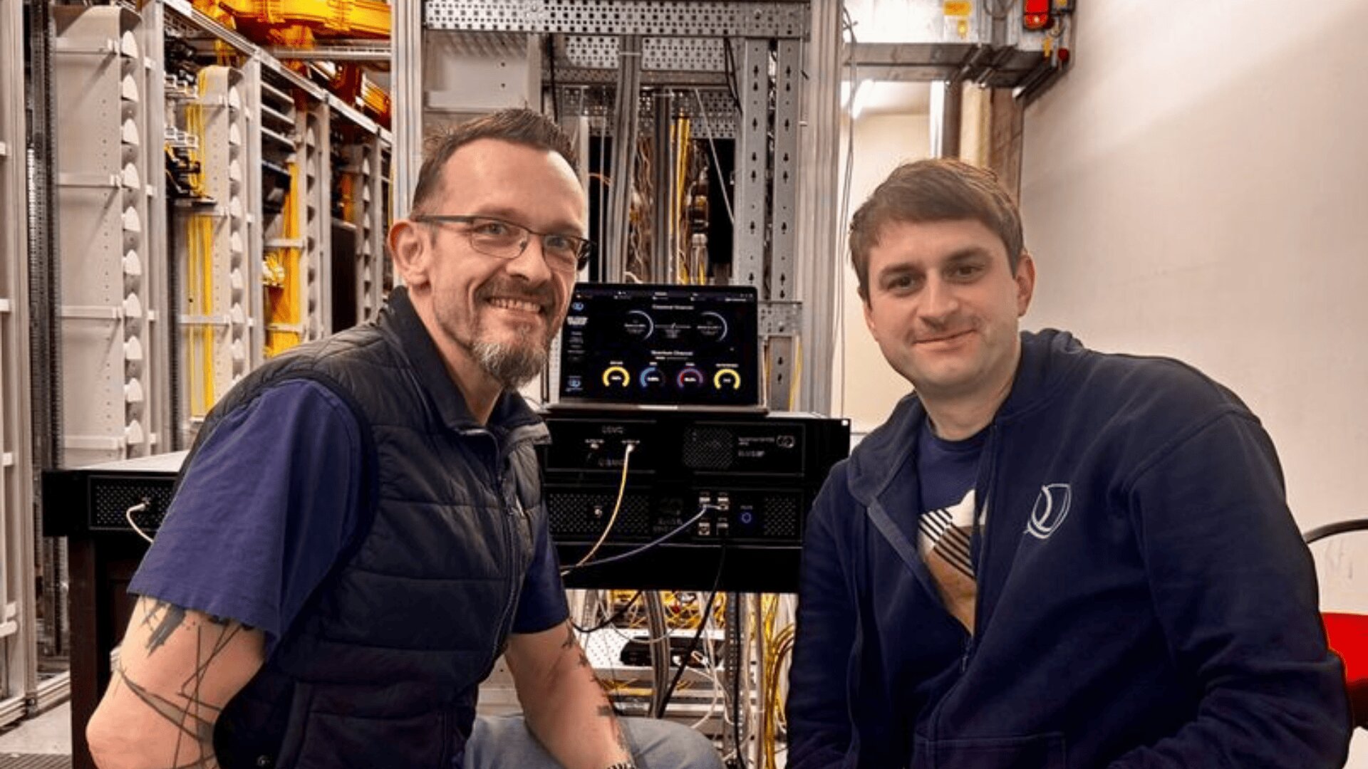 Zwei Männer sitzen lächelnd vor Computergeräten in einem Serverraum. Dahinter sind Kabel und elektronische Geräte zu sehen. Einer trägt eine Brille und eine Weste, der andere einen dunkelblauen Kapuzenpullover.