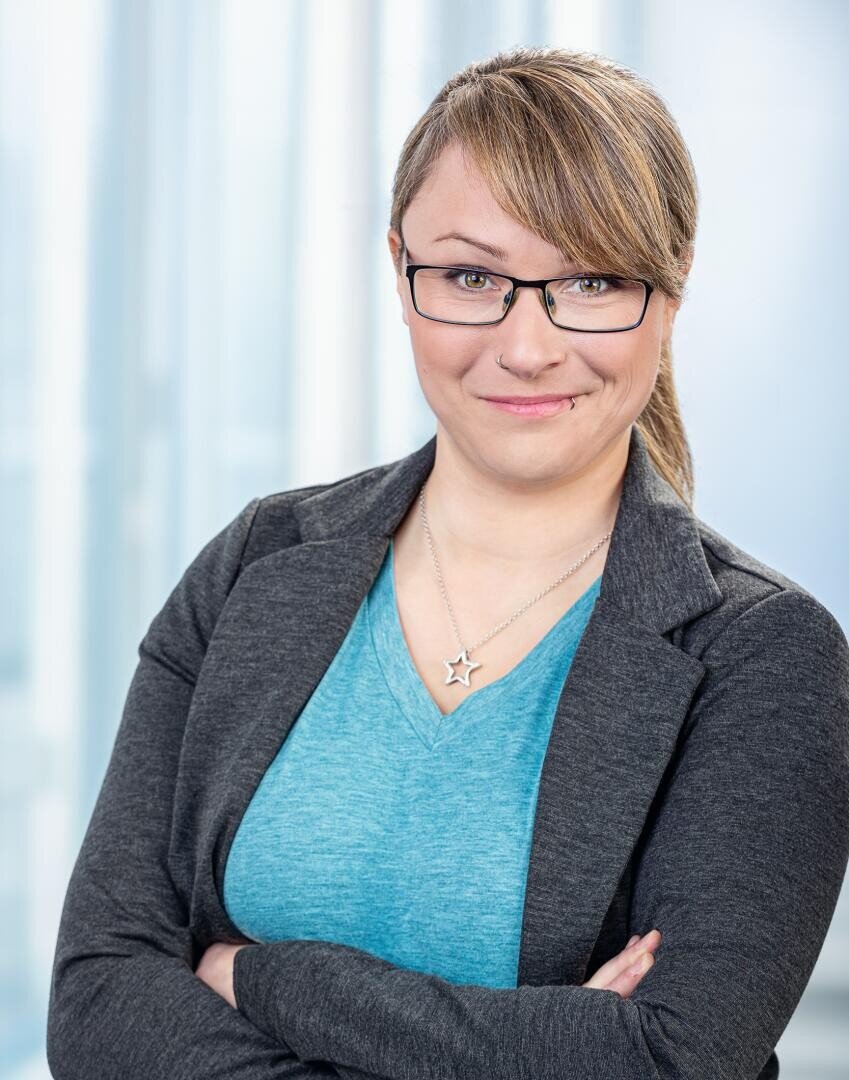 Eine Frau mit hellbraunem Haar zu einem Pferdeschwanz, Brille und einem grauen Blazer über einem blauen Oberteil steht mit verschränkten Armen da und lächelt leicht. Sie trägt eine Halskette mit Sternen. Der Hintergrund ist sanft in hellblauen Tönen verwischt.
