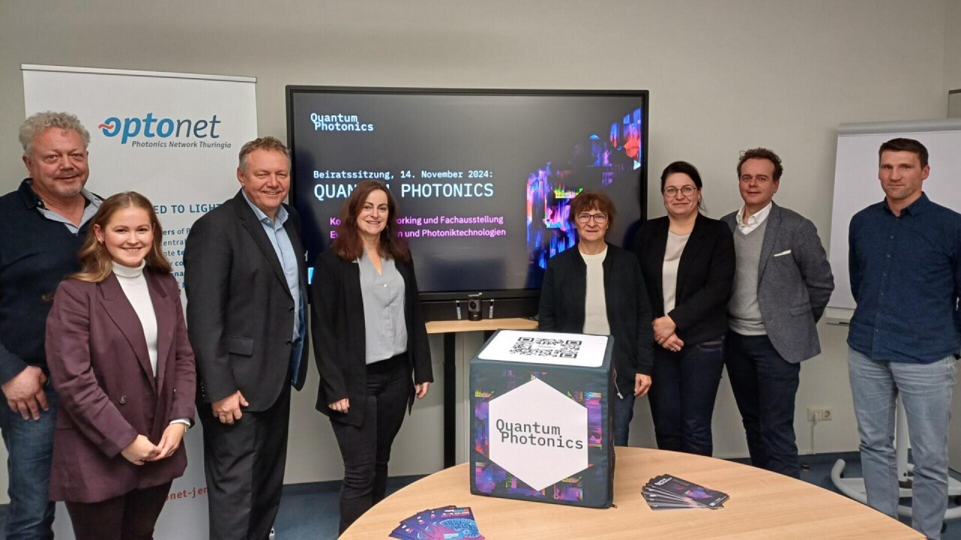Eight people stand smiling in front of a screen displaying “Quantum Photonics”; brochures and a cube with the event logo are on a round table; banners and office setting visible in the background.