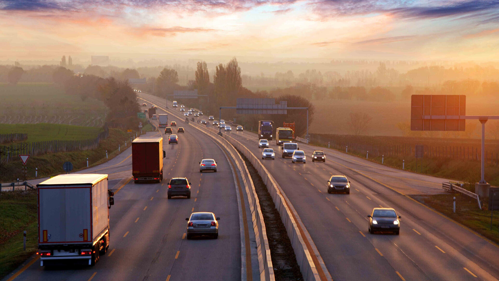 Autos und Lastwagen fahren bei Sonnenaufgang oder Sonnenuntergang auf einer geteilten Autobahn durch eine ländliche Landschaft, mit wenig Verkehr in beide Richtungen und einem bunten Himmel über ihnen.