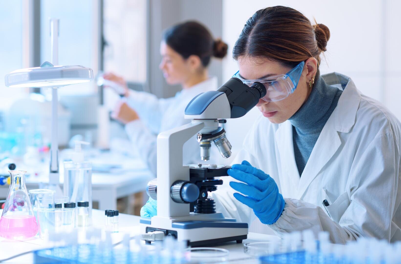 Ein Wissenschaftler mit Schutzbrille und Handschuhen untersucht in einem Labor Proben unter einem Mikroskop. Ein weiterer Wissenschaftler arbeitet im Hintergrund. Auf den Labortischen sind Laborgeräte und Glaswaren zu sehen.