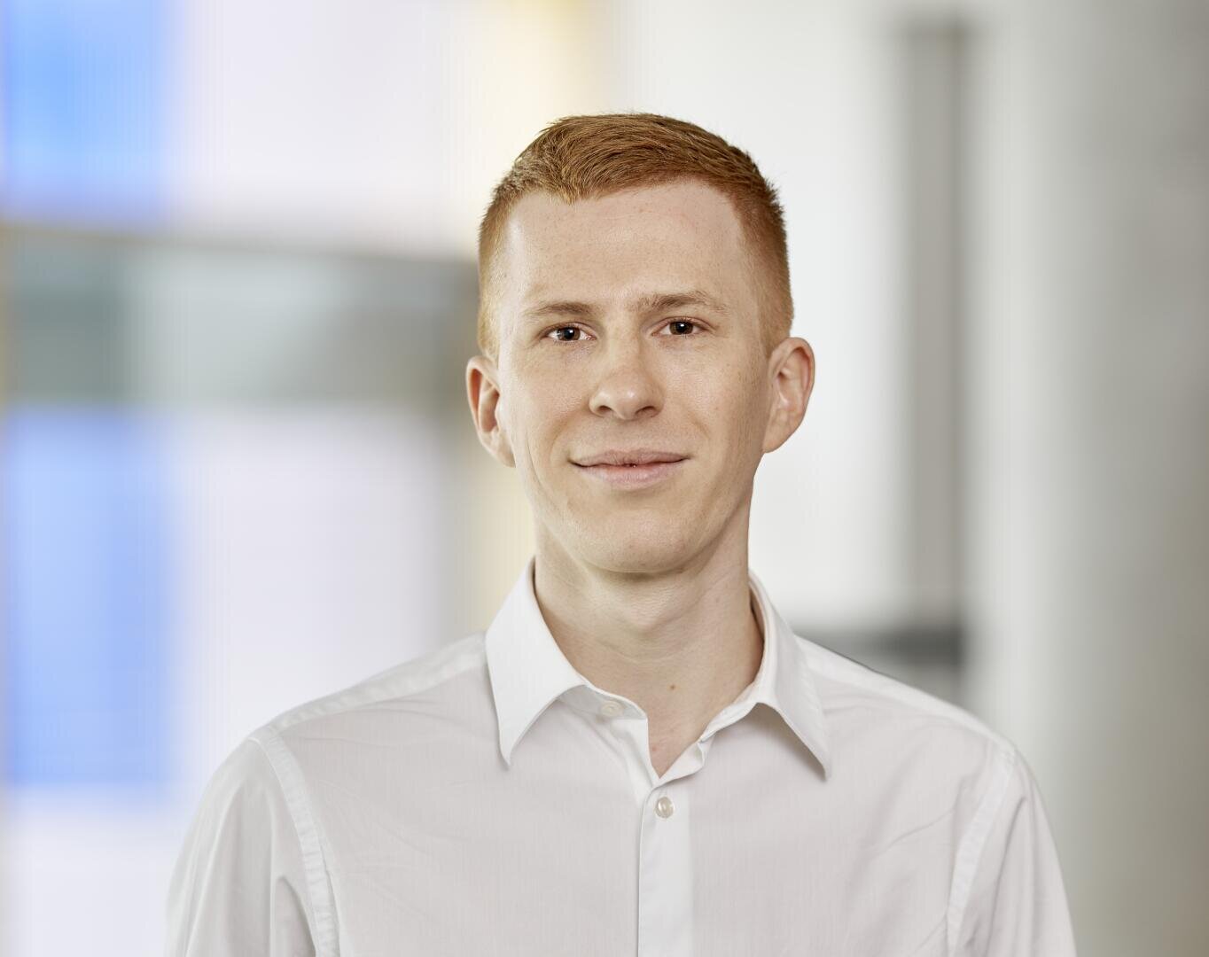 Eine junge Person mit kurzen roten Haaren, heller Haut und einem leichten Lächeln, die ein weißes Hemd mit Knöpfen trägt, steht vor einem leicht verschwommenen Innenhintergrund.