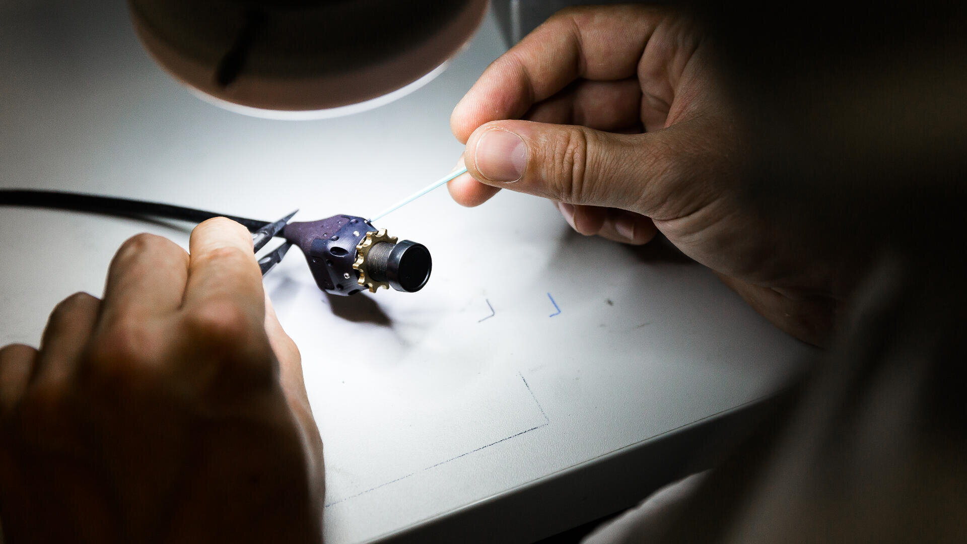 Die Hände einer Person arbeiten unter einer Lampe und montieren oder reparieren mithilfe einer Pinzette oder eines Werkzeugs auf einer weißen Arbeitsfläche ein kleines elektronisches Gerät.