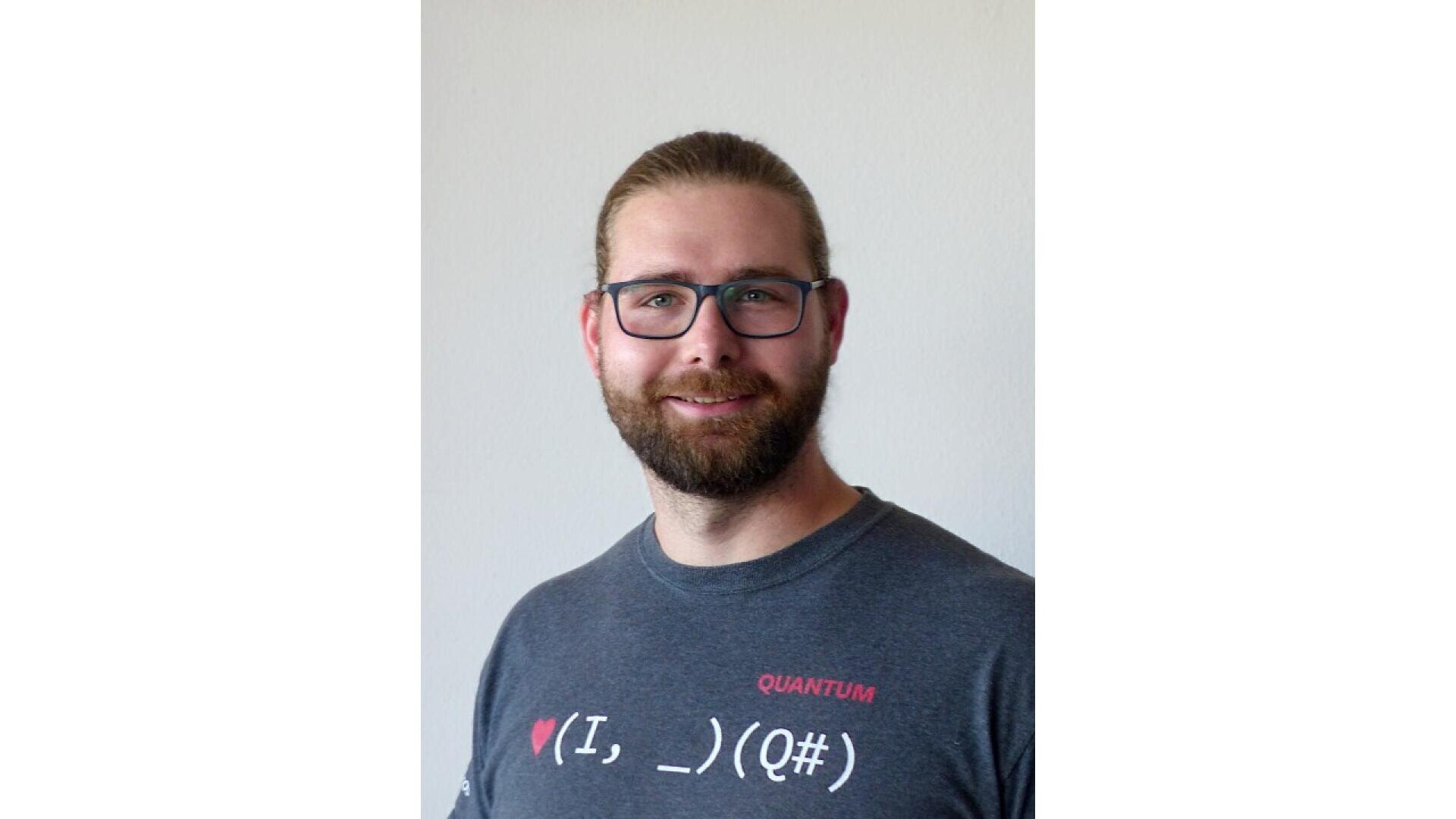 A man with glasses and a beard smiles at the camera. He is wearing a dark gray T-shirt with the word QUANTUM and a math-themed graphic on it. The background is plain and light-colored.
