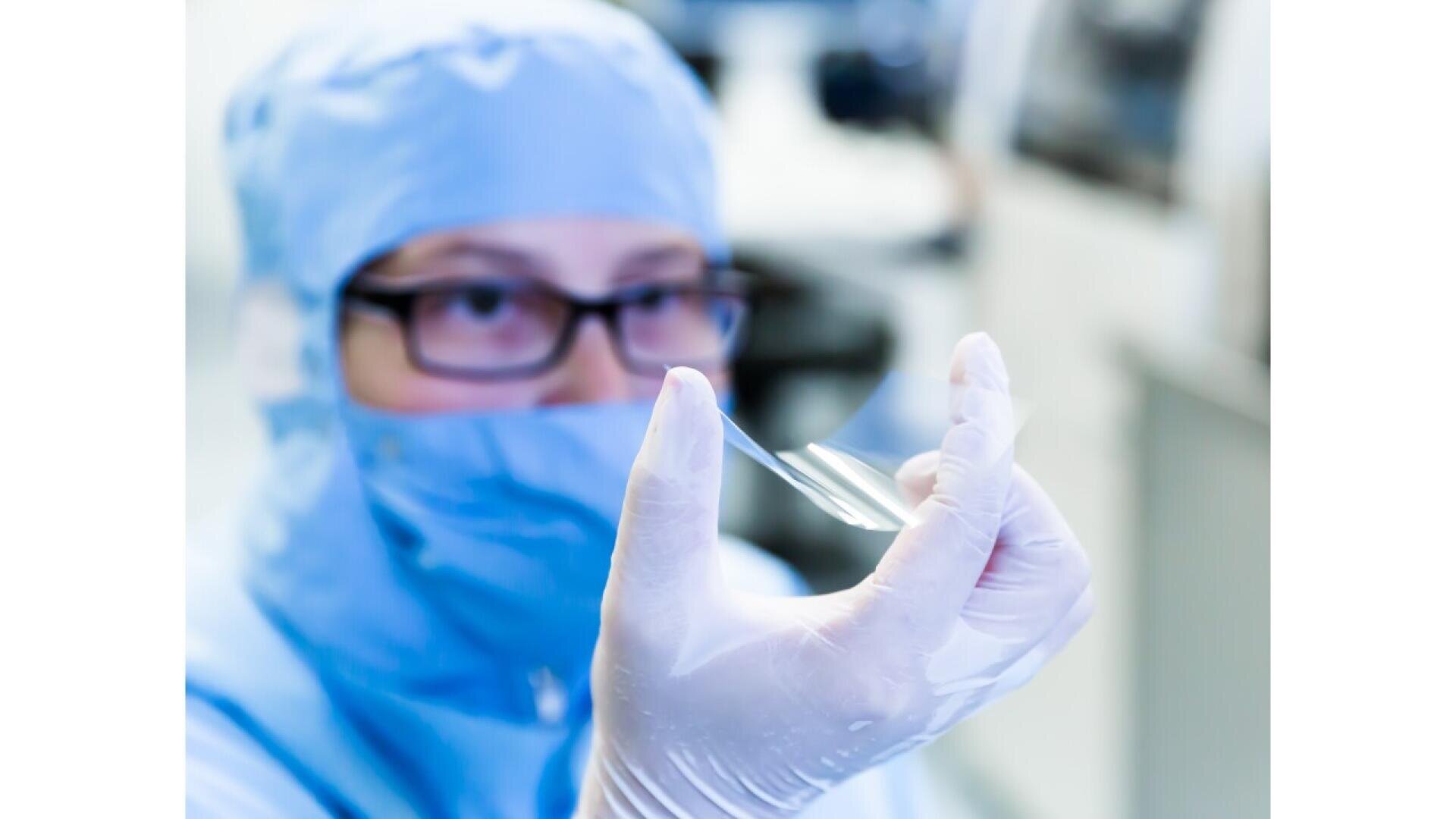 A scientist wearing protective gear and gloves examines a transparent, thin material in a laboratory setting. The focus is on the sample, with lab equipment blurred in the background.