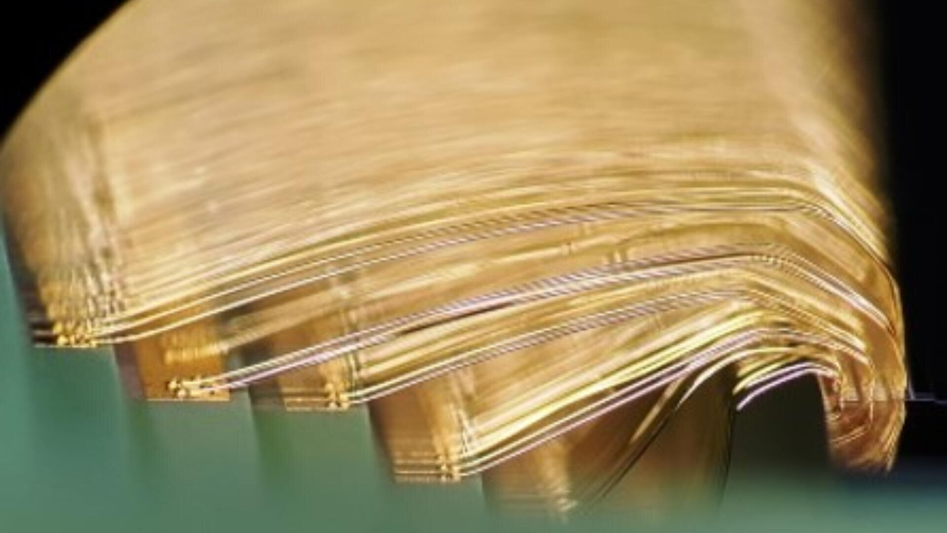 Eine Nahaufnahme dünner, flexibler, goldfarbener Metallplatten oder -drähte, die geschichtet und leicht gebogen sind, das Licht reflektieren und vor einem dunklen Hintergrund ein leuchtendes, abstraktes Erscheinungsbild erzeugen.