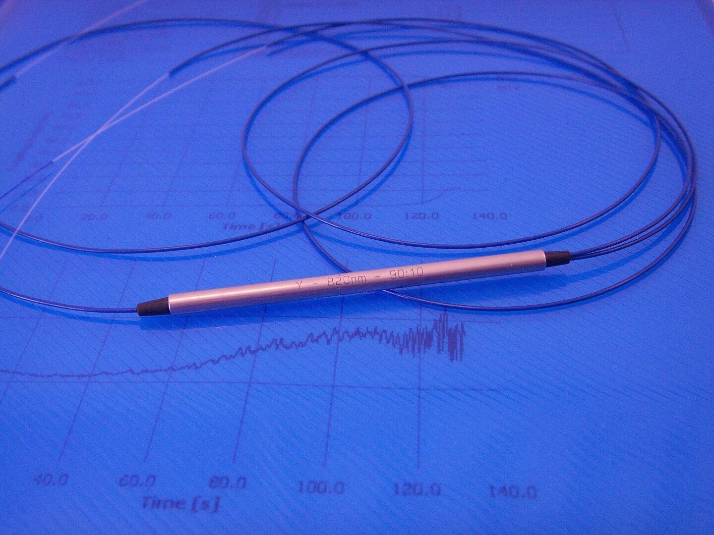 A slim metallic fiber optic sensor with thin blue cables is placed on a blue surface that has scientific graphs and data, including a plot labeled “Time [s].”.