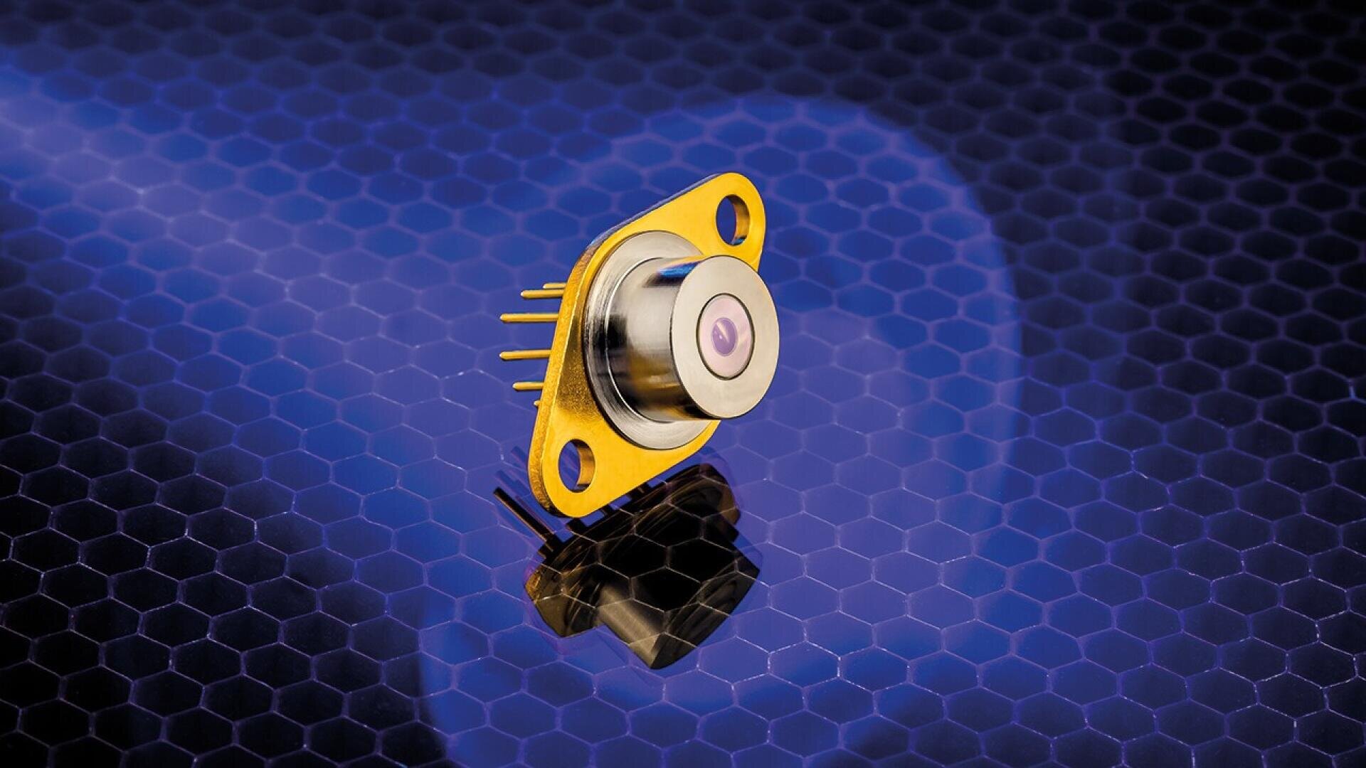 A gold-colored electronic component with a round center and two screw holes sits on a blue, hexagonal-patterned surface, casting a reflection below. Several gold pins extend from its base on one side.
