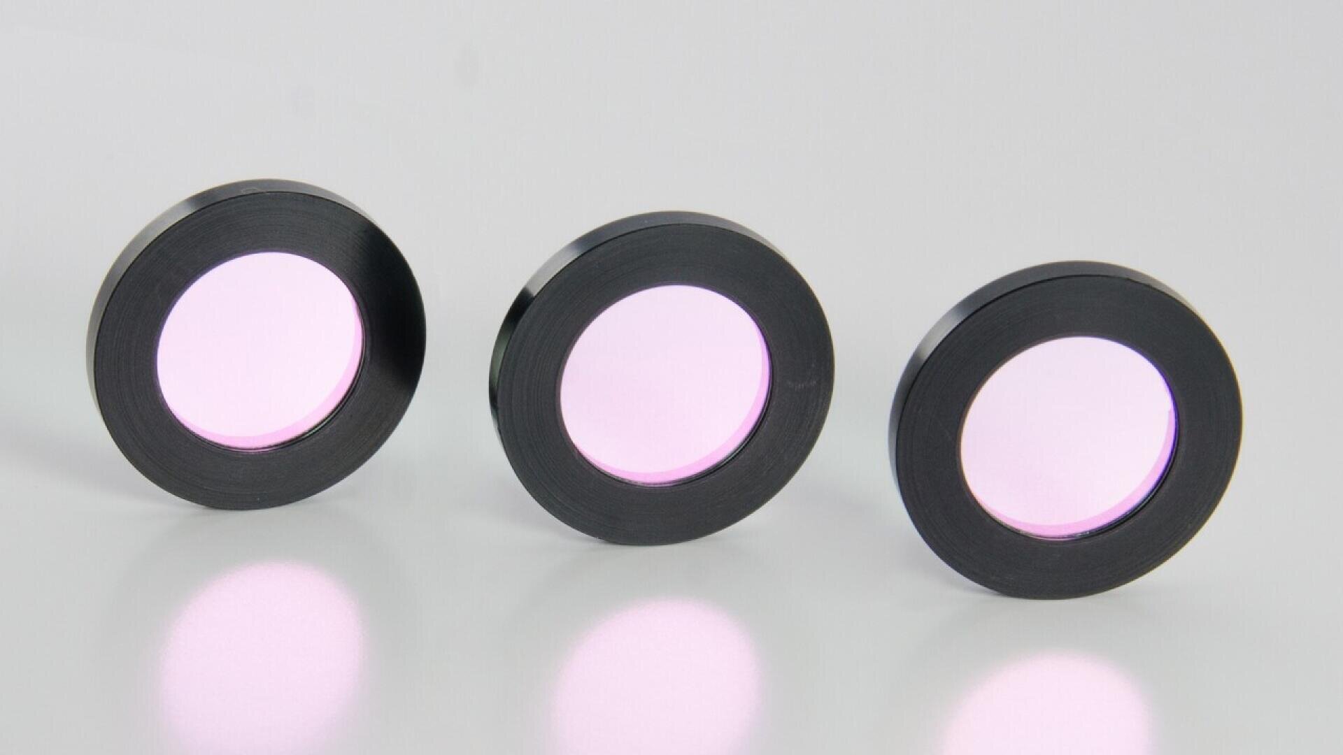 Drei runde optische Filter mit schwarzen Außenrändern und hellviolett getönten Glaszentren stehen aufrecht auf einer reflektierenden weißen Oberfläche.