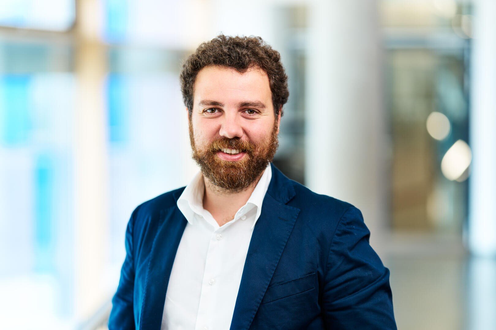 Ein Mann mit lockigem braunem Haar und Bart, der ein weißes Hemd und einen dunkelblauen Blazer trägt, lächelt in einer hellen, modernen Inneneinrichtung mit unscharfen Hintergrundelementen in die Kamera.