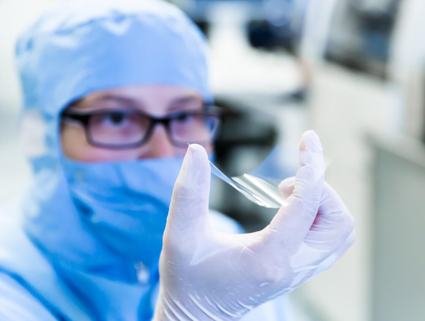 Ein Wissenschaftler in Schutzkleidung und Handschuhen untersucht in einer Laborumgebung eine dünne, transparente Folie. Der Hintergrund ist unscharf, wodurch der Fokus auf die Folie und die aufmerksame Beobachtung des Wissenschaftlers gerichtet ist.