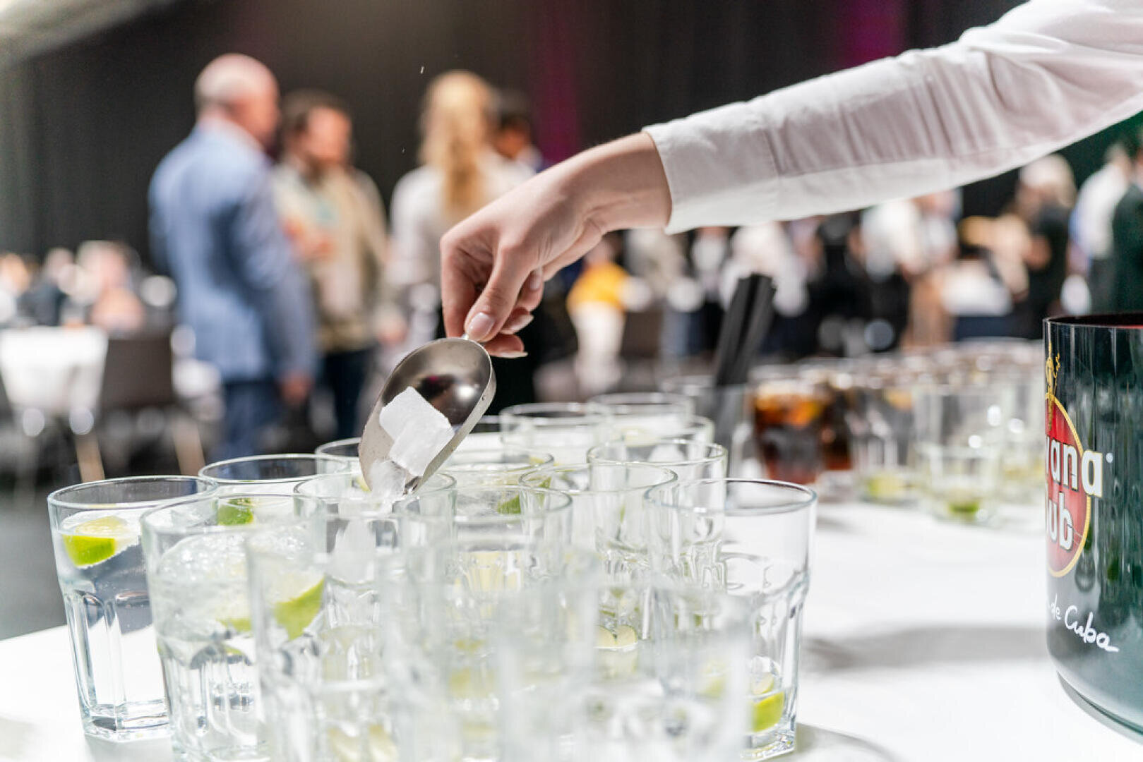 Die Hand einer Person gibt einen Eiswürfel in ein Glas mit Limettenscheiben auf einem mit ähnlichen Gläsern gefüllten Tisch, während sich im unscharfen Hintergrund bei einer Veranstaltung in geschlossenen Räumen Menschen versammeln und unterhalten.