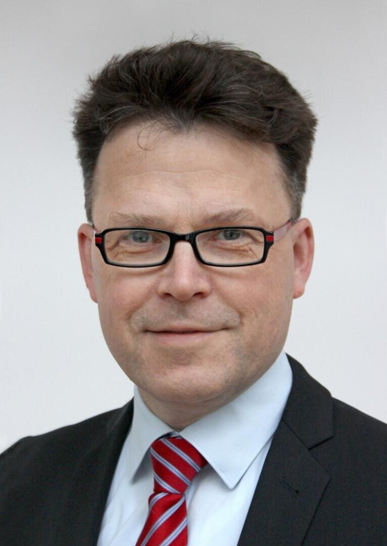 Ein Mann mit Brille, dunklem Anzug, hellblauem Hemd und rot gestreifter Krawatte, leicht lächelnd, mit kurzen dunklen Haaren und neutralem Hintergrund.