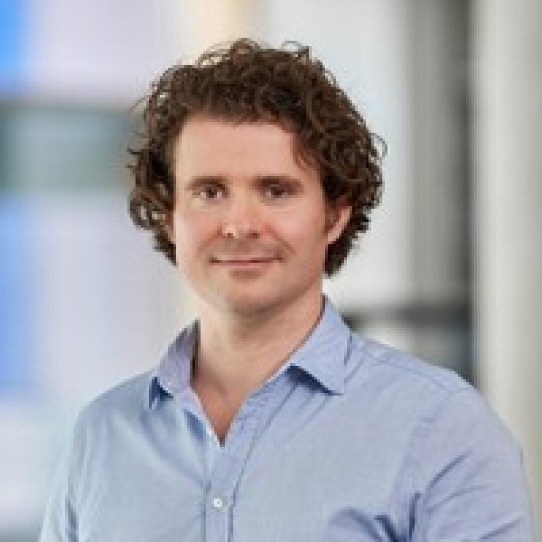 Ein Mann mit lockigem braunem Haar und einem hellblauen Hemd mit Kragen steht in einem Innenbereich vor einem unscharfen Hintergrund mit sanftem Tageslicht.