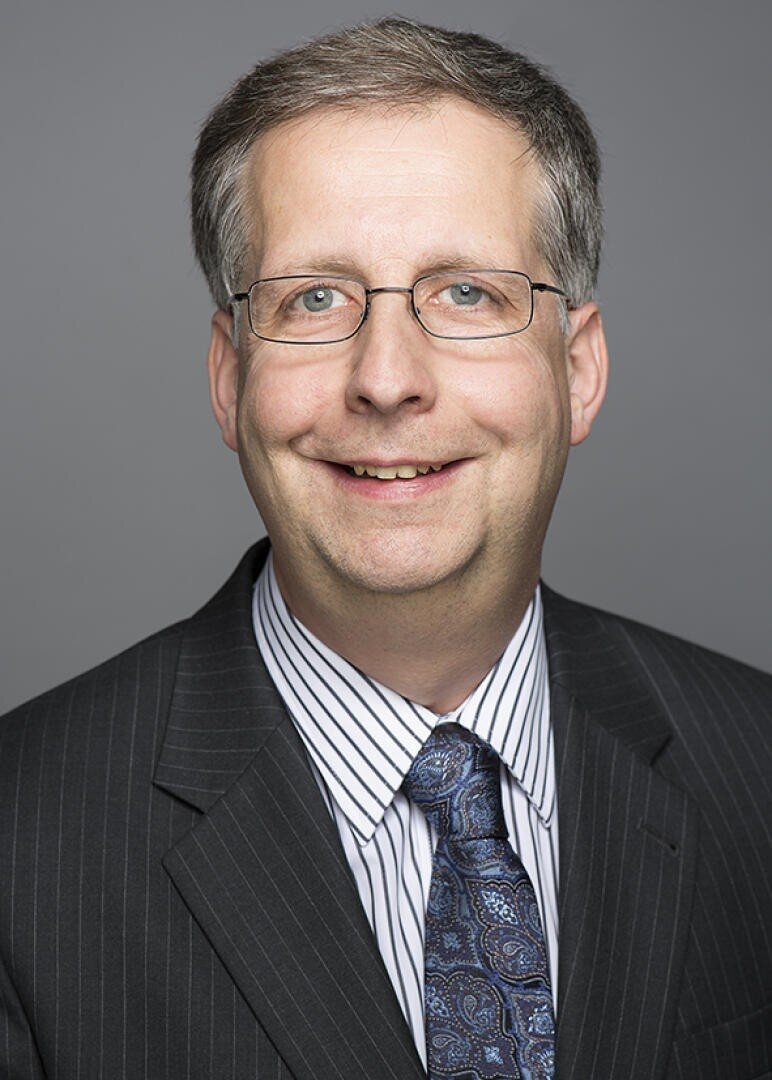 Ein Mann mittleren Alters mit kurzen grauen Haaren, Brille und einem gestreiften Jackett über einem weißen Hemd mit dünnen Streifen und einer gemusterten blauen Krawatte, der vor einem schlichten grauen Hintergrund lächelt.