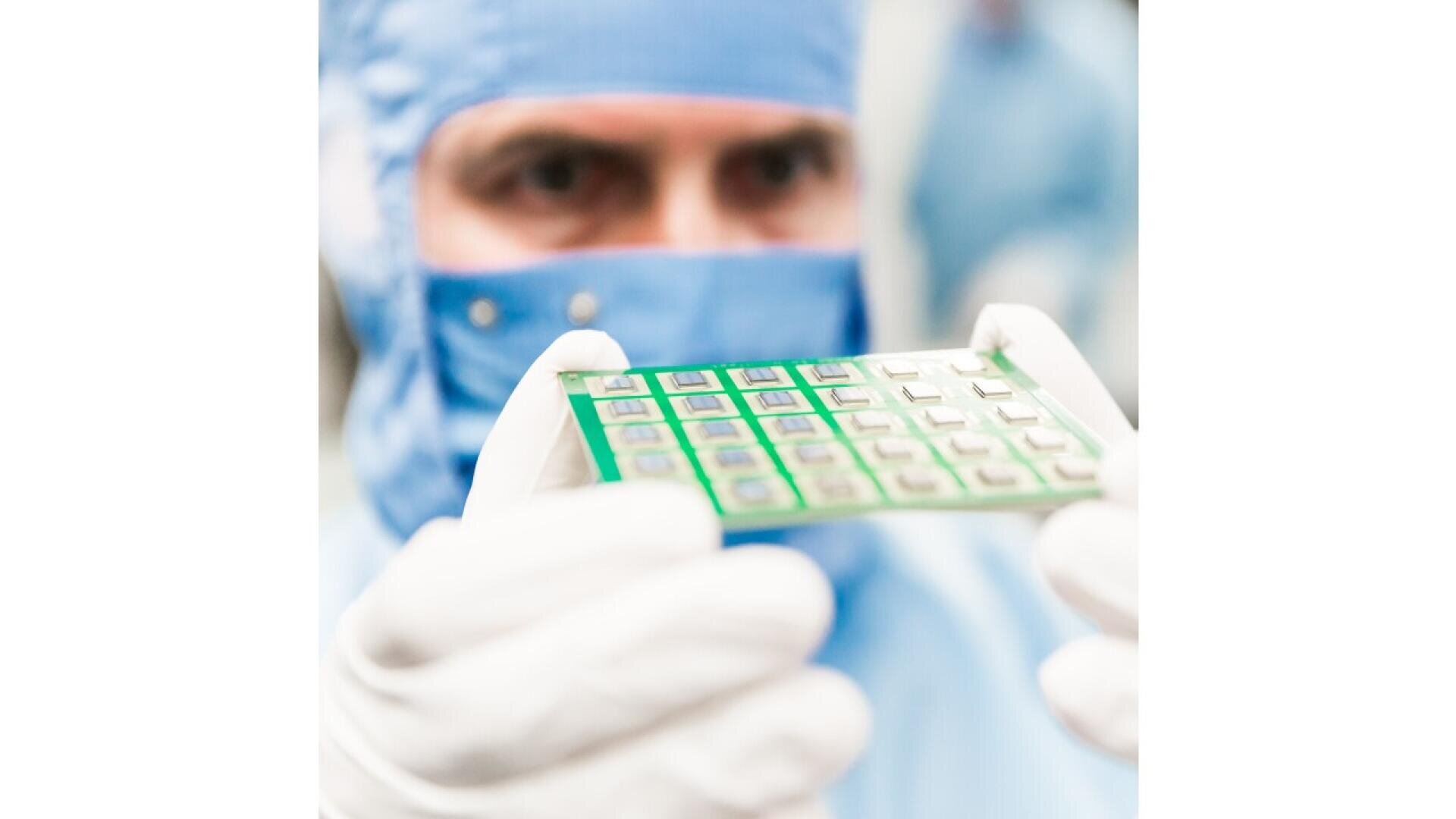 A person in protective clothing and gloves holds a green microchip or circuit board, examining it closely in a clean, laboratory-like environment.
