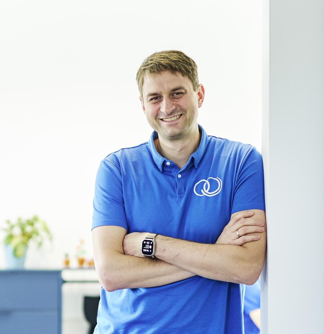 Ein Mann in einem blauen Poloshirt mit weißem Logo steht lächelnd mit verschränkten Armen an einer weißen Wand in einem hellen Raum mit einer Pflanze und Möbeln im Hintergrund.