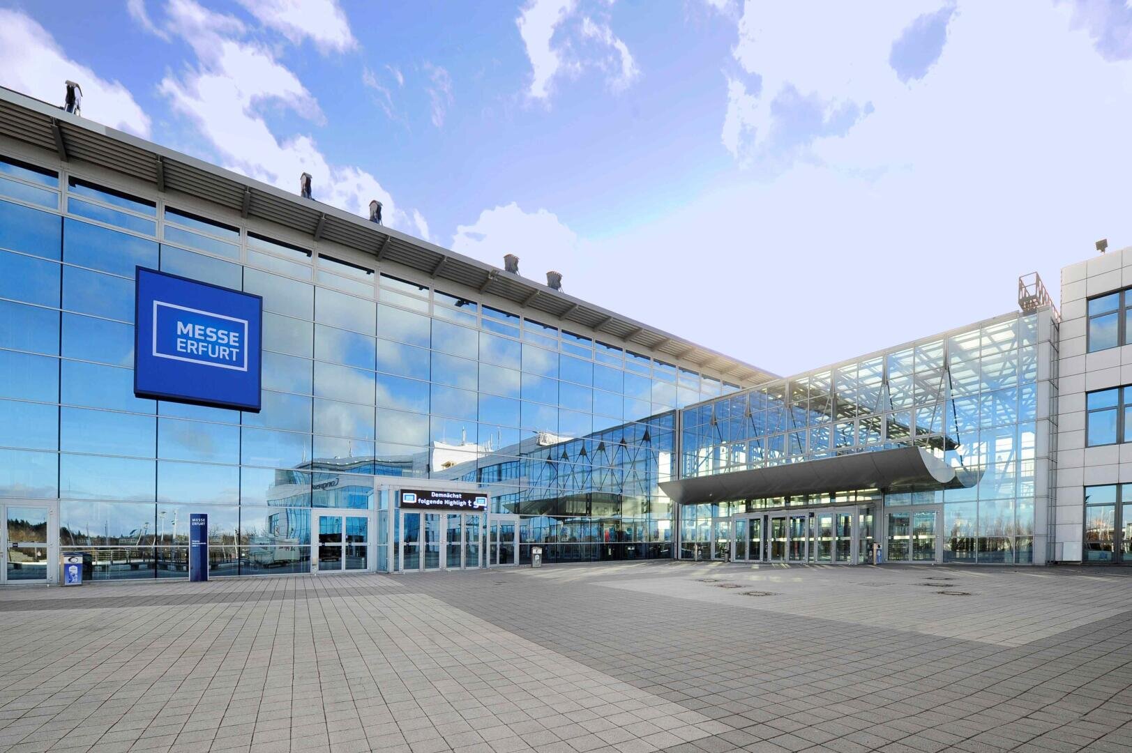 Das Bild zeigt die Außenansicht der Messe Erfurt, einem modernen Kongresszentrum mit großen Glasfenstern, in denen sich der Himmel spiegelt, einem markanten blauen Schild und mehreren Eingangstüren zu einem geräumigen gepflasterten Platz.