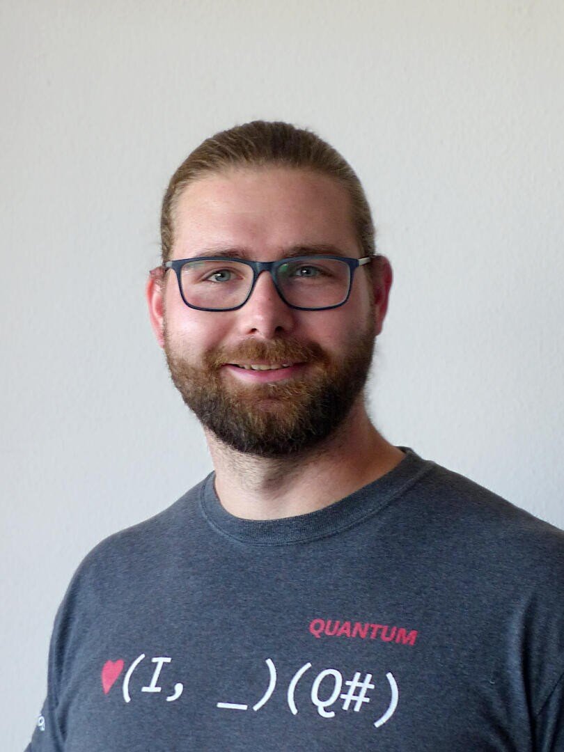 Ein Mann mit Bart und Brille lächelt in die Kamera. Er trägt ein dunkles T-Shirt mit dem Schriftzug „QUANTUM“ und einer Programmiergrafik. Der Hintergrund ist schlicht und hell.
