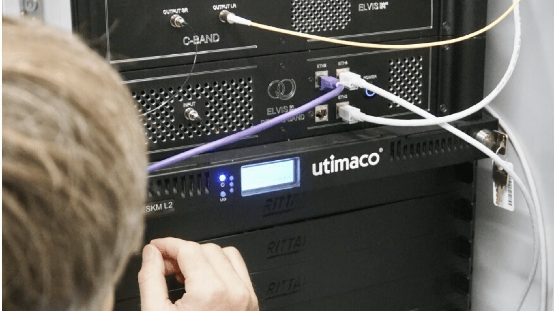 A person adjusts cables connected to a rack-mounted Ultimaco device in a server room, with various network cables and hardware components visible.