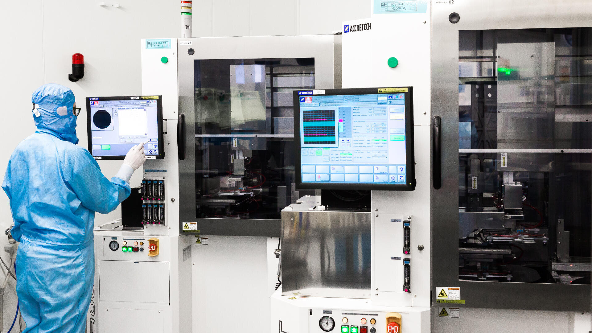 Eine Person in blauer Schutzkleidung bedient ein Touchscreen-Panel in einem Reinraumlabor mit modernen Maschinen und Monitoren, die Daten und Systemsteuerungen anzeigen.