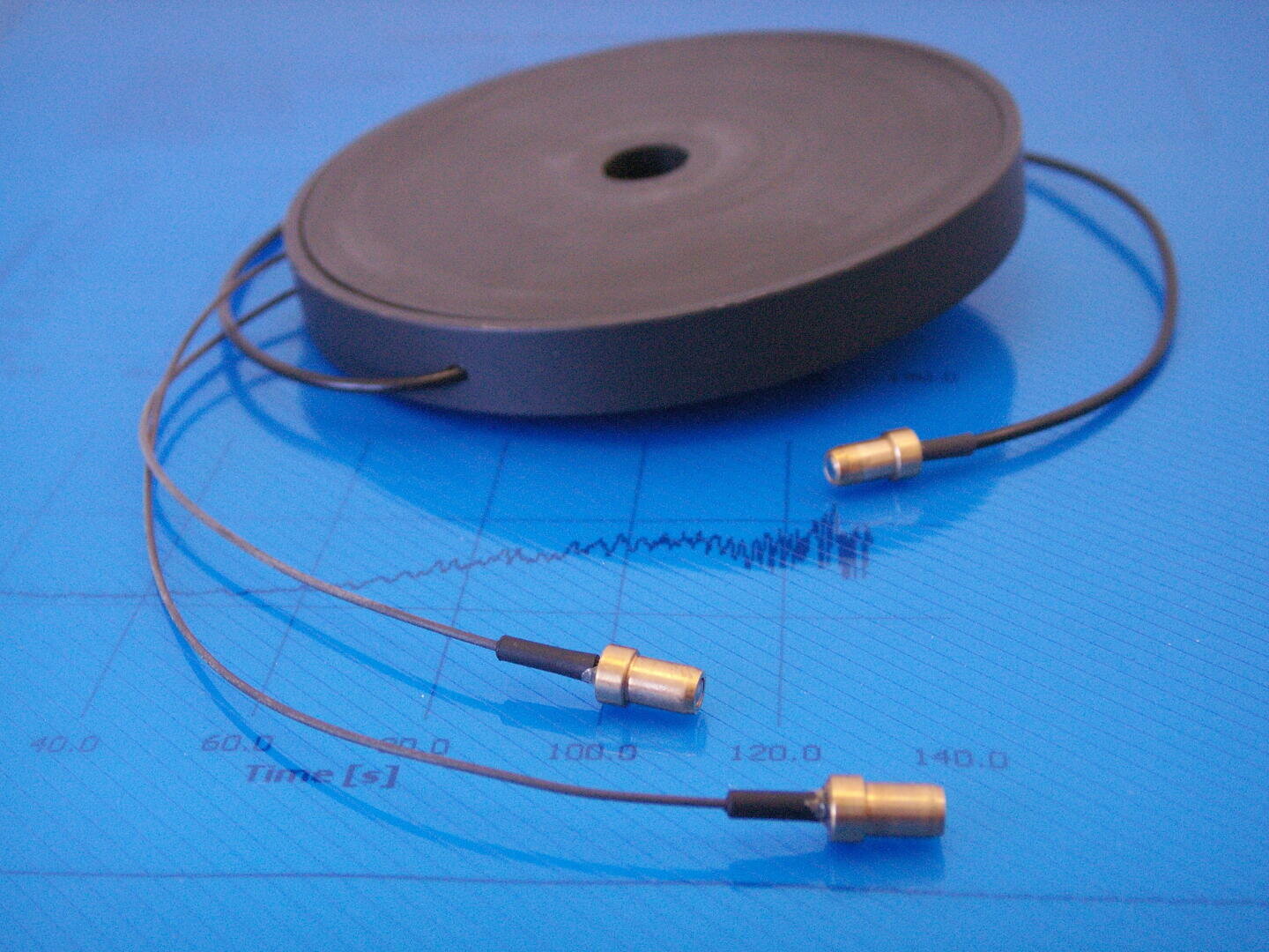 A round black coil with three attached cables ending in gold connectors is placed on a blue surface with faint wave graphs and time measurements in the background.