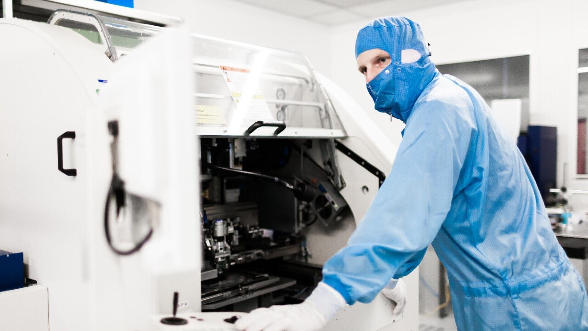 A person in a blue cleanroom suit and mask operates industrial equipment in a bright, sterile laboratory or manufacturing facility.