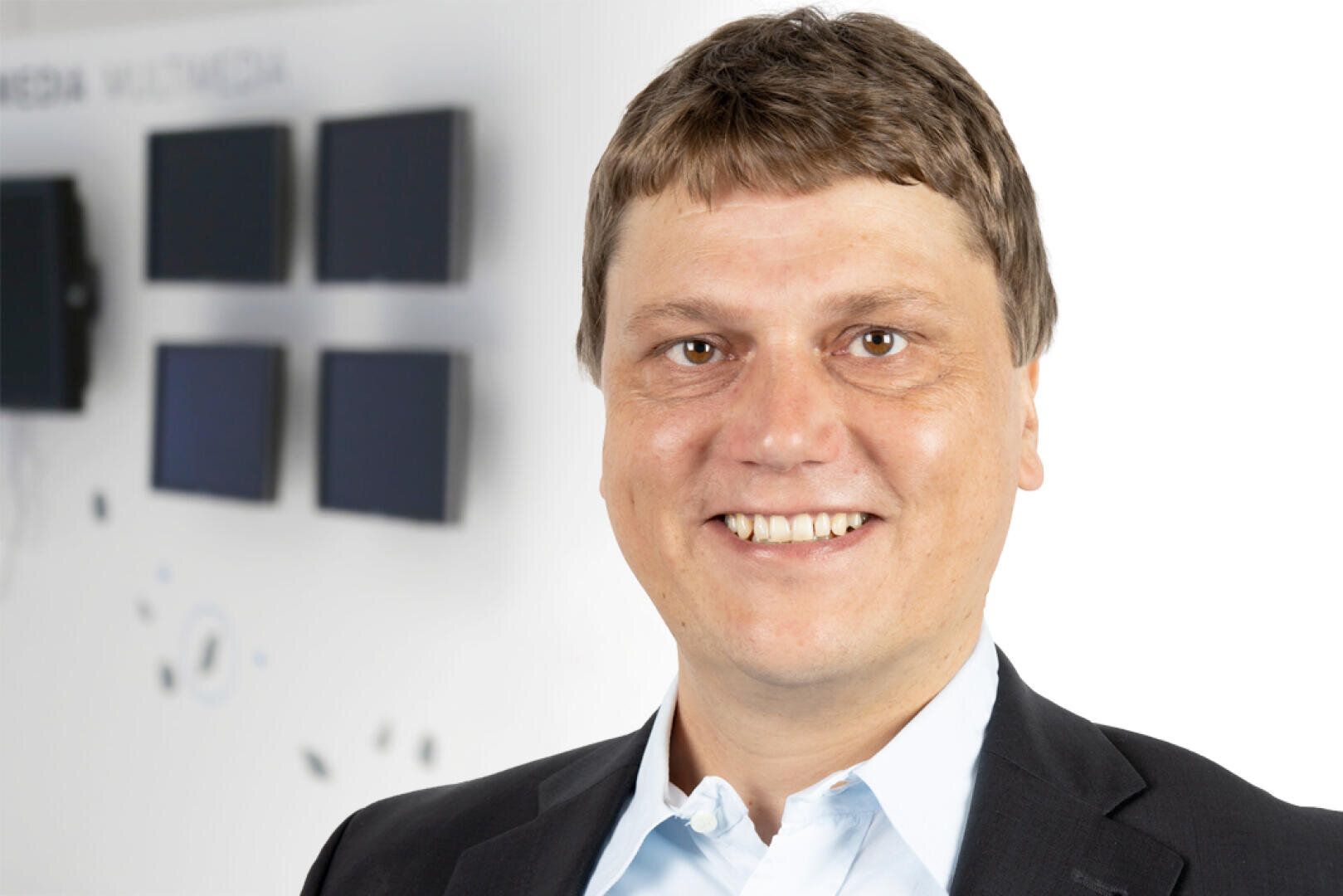 Ein lächelnder Mann im Jackett und hellblauem Hemd steht vor einer unscharfen Wand mit kleinen schwarzen Bildschirmen und schwachem Text im Hintergrund.