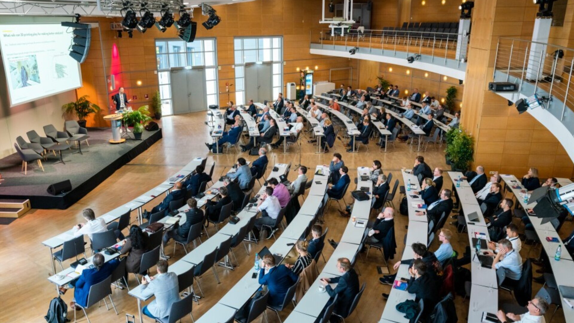 A large conference room with tiered seating, filled with attendees facing a stage where a presentation is being shown on a screen. The room has wood-paneled walls and modern design elements.