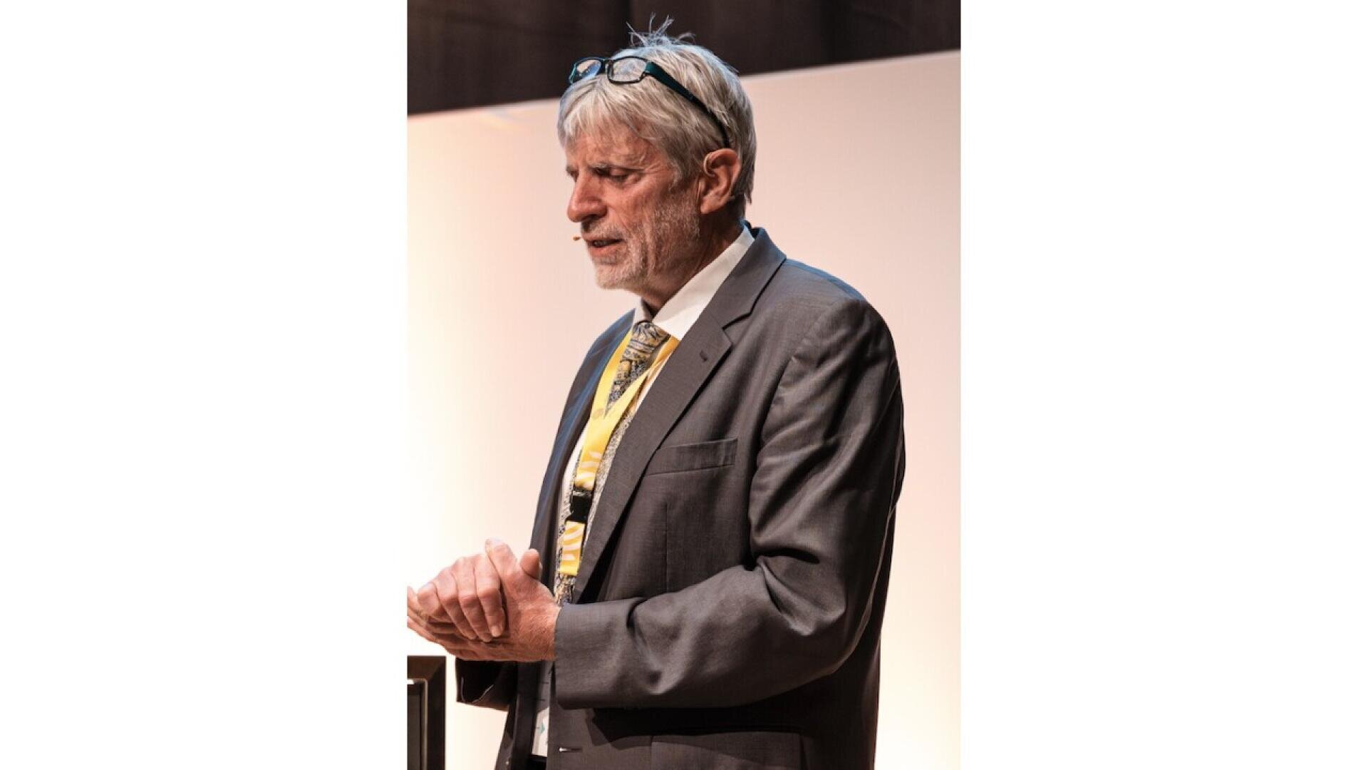 A man in a suit and tie stands indoors, speaking with his hands clasped. He has gray hair and glasses resting on his head.