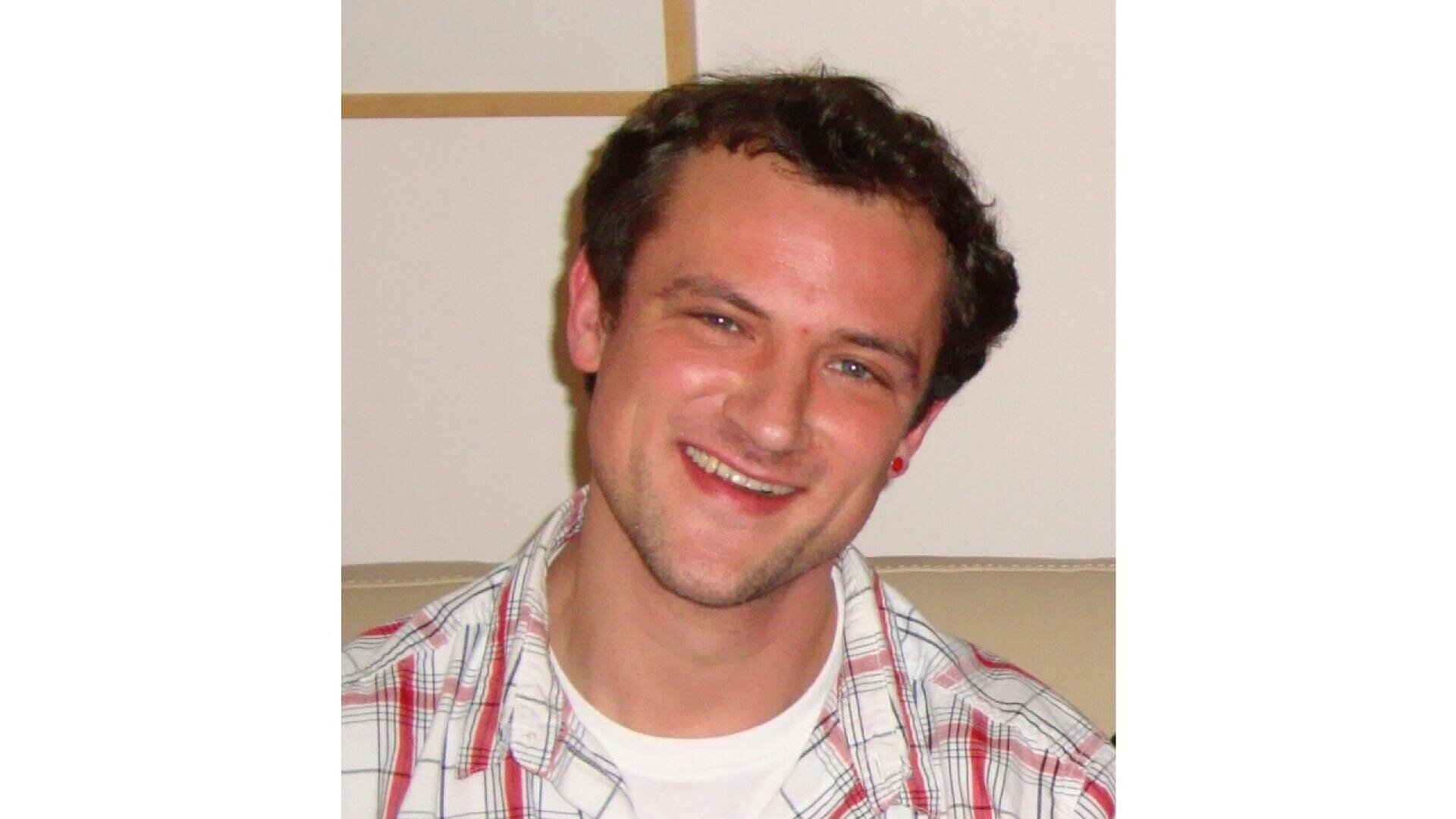 A man with short brown hair is smiling at the camera. He is wearing a white T-shirt under a red and white plaid shirt, sitting indoors against a light-colored background.