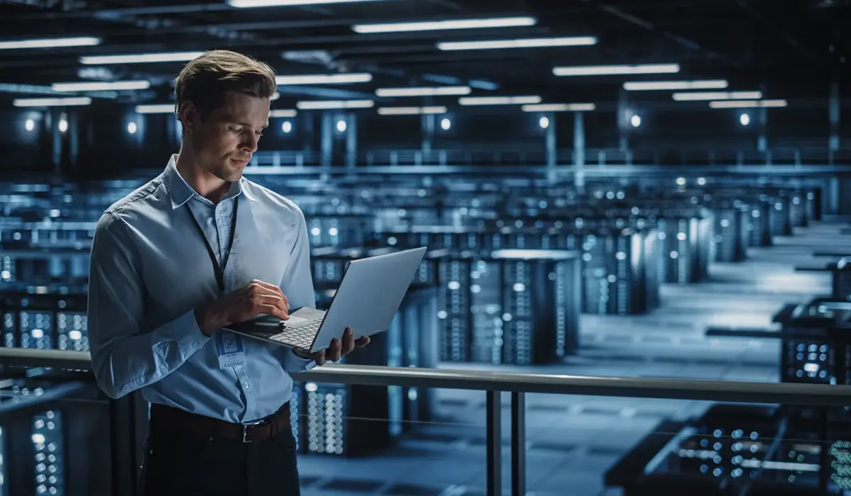 Ein Mann mit Laptop steht auf einer Galerie über einem großen Raum voller Serverracks und moderner Computertechnik.